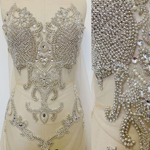 May include: A beige fabric adorned with an elaborate, symmetrical design of sparkling rhinestones and faux pearls. The embellishment is densely packed, creating a shimmering effect, ideal for bridal or formal wear.