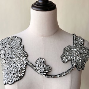 Op de afbeelding: Een zilveren kralenversiering voor kleding, met een bloemmotief en een centrale cluster van kralen. De versiering is op een zwarte mesh-rug.