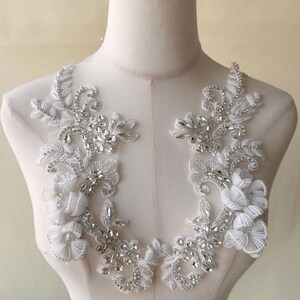 May include: A pair of white beaded lace appliques featuring silver rhinestones and pearl-like embellishments. The appliques are designed in a floral pattern with delicate branches and leaves.