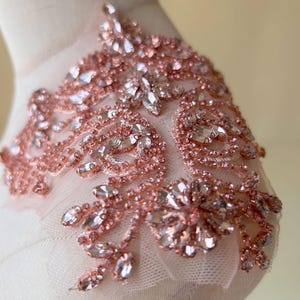 May include: A rose gold applique featuring intricate floral patterns crafted from clear faceted crystals and small rose gold beads. The decorative piece is set on a sheer, light-colored mesh fabric, showcasing a detailed arrangement of marquise and round stones.
