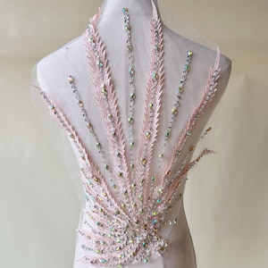 Puede incluir: Una aplicación rosa con cuentas y lentejuelas con pedrería iridiscente. La aplicación tiene forma de abanico y presenta un diseño delicado y femenino.