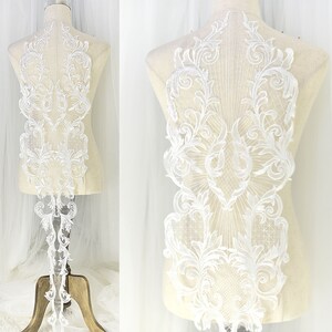 May include: White floral lace applique with an intricate design. The lace features a symmetrical pattern of swirling leaves and floral motifs. The applique is displayed on a mannequin, showcasing its shape and detail. Perfect for bridal wear or other decorative projects.