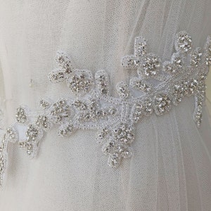 May include: White tulle fabric with a silver rhinestone trim. The trim features a floral design with leaves and flowers.