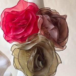 May include: Three large decorative flower brooches made of layered, translucent organza fabric. The flowers are colored in vibrant fuchsia, muted mauve, and soft gold, each featuring delicate, ruffled petals with finished edges.