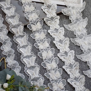 May include: White lace trim with a delicate butterfly design. The trim features a 3D effect with pearl embellishments.