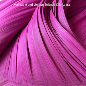 Puede incluir: Una tela de color magenta vibrante con una textura estriada distintiva y una apariencia fluida y drapeada. El material lleva el texto Malleable and Unique Striated Silk Abaca, mostrando su composición flexible y en capas.