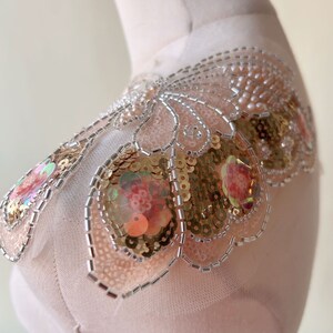 May include: A shoulder applique with a floral design. It features gold and iridescent sequins, and silver beads. The base is a sheer, pale pink fabric. This decorative piece would be suitable for embellishing clothing or accessories.