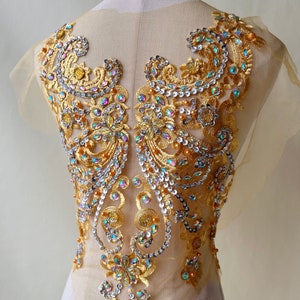 Puede incluir: Aplique de cuentas doradas e iridiscentes con un diseño floral. El aplique está sobre un fondo de tela transparente.