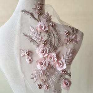 May include: A delicate floral applique featuring pink 3D flowers, leaves, and pearl beads. The applique is ideal for adding a touch of elegance to clothing, accessories, or home decor.