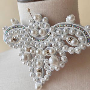 Peut inclure: Un collier ras de cou blanc orné de perles et de strass. Le collier est fait de tissu blanc avec un délicat motif floral. Les perles sont disposées en grappes et sont rehaussées de petits strass.