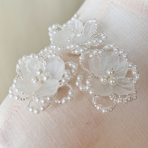 Peut inclure: Trois fleurs blanches en perles avec des accents de perles. Les fleurs sont faites d'un tissu blanc délicat et sont entourées d'une bordure perlée.