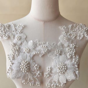 Könnte beinhalten: Weiße florale Spitzenapplikation mit Perlenakzenten. Die Applikation zeigt detaillierte florale Designs mit Clustern aus kleinen Perlen. Die Spitze ist zart und aufwendig, geeignet zum Verzieren von Kleidungsstücken oder Bastelarbeiten. Das Design ist symmetrisch und elegant.