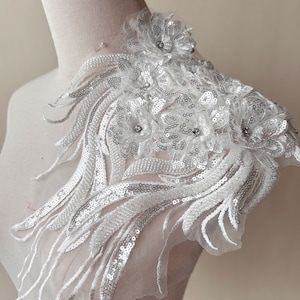 May include: White lace applique with silver sequins and rhinestones. The applique features a floral design with delicate feathers. This applique is perfect for adding a touch of elegance to any garment.