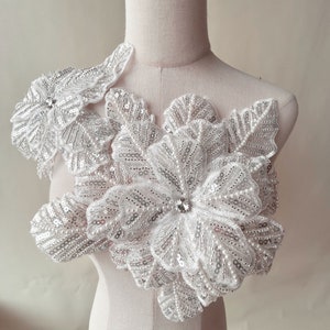 May include: White floral applique with pearl and sequin embellishments. The applique is made of delicate fabric and features a layered design.