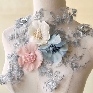 May include: A floral applique featuring three fabric flowers in white, light blue, and pink, with silver embroidered detailing. The flowers are attached to a sheer, silver-embroidered base, creating a delicate and elegant design. Ideal for embellishing garments or accessories.
