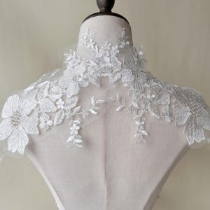 Peut inclure: Une applique en dentelle blanche avec des broderies argentées, présentant des motifs floraux. L'applique est présentée sur un mannequin blanc.