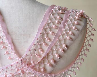 Könnte beinhalten: Ein filigraner, rosafarbener Perlenbesatz mit netzartigem Design und herabhängenden perlenartigen Perlen. Der Besatz hat ein transparentes rosa Band und wird auf einer Schaufensterpuppe gezeigt. Der Besatz dient wahrscheinlich zum Verzieren von Kleidung oder Accessoires.