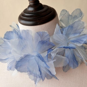 May include: A delicate blue and white flower collar made of layered fabric. The collar is made of soft, sheer fabric and has a ruffled, layered design.