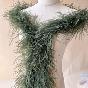 Op de afbeelding: Een saliegroene veren boa gedrapeerd over een witte etalagepop. Het accessoire bestaat uit lange, warrige, natuurlijk ogende veren in een gedempte olijfgroene tint, wat zorgt voor een volumineus en getextureerd uiterlijk rond de hals en borst.