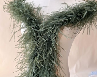 May include: A long, sage green feather boa made of soft, wispy plumes. The boa is draped around the neck and shoulders of a white mannequin, showcasing the dense, layered texture of the individual feathers that create a full, voluminous appearance.