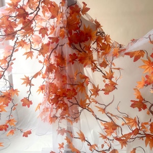 May include: A delicate white fabric featuring orange and red maple leaves and dark brown branches. The leaves vary in size and are spread across the material, creating an autumnal design. The fabric is likely intended for decorative use, such as for a veil.