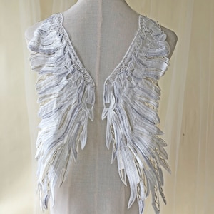 May include: White lace applique featuring a wing design. The lace is delicate and has a scalloped edge. The wings are symmetrical and have a flowing, ethereal look.