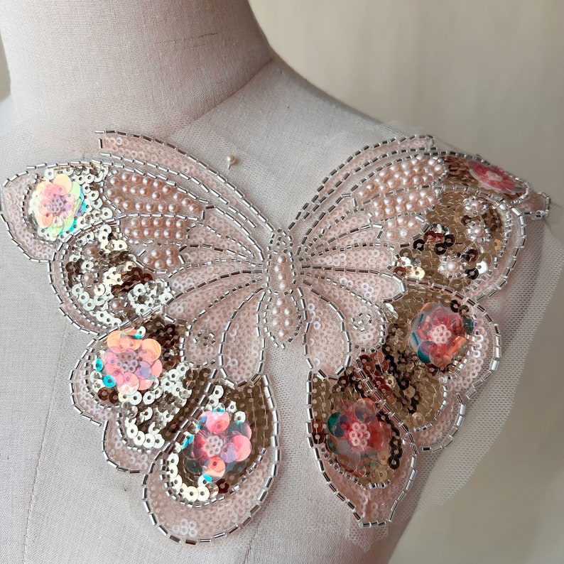 May include: A decorative butterfly applique with a light pink base, embellished with sequins, beads, and small floral accents. The butterfly design features a detailed outline and intricate patterns, creating a delicate and ornate appearance.