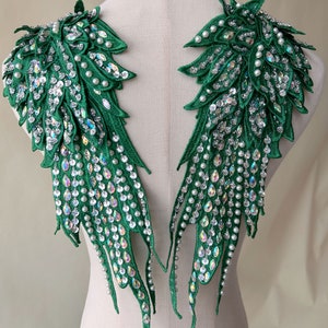 Può includere: Un paio di ali in tessuto verde con paillettes iridescenti e perline di perle bianche. Le ali sono progettate per essere cucite su un capo di abbigliamento.