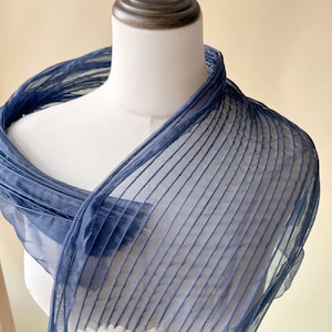 Könnte beinhalten: Ein dunkelblauer, transparenter Schal mit einer fein plissierten Textur und vertikalen Linien im gesamten Stoff. Das leichte Material ist um einen Schaufensterpuppenhals drapiert und zeigt die halbtransparente Beschaffenheit sowie das gleichmäßige Rippenmuster.