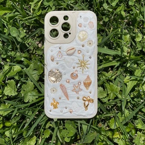 May include: A white iPhone case decorated with seashells, starfish, pearls, and gold accents. The case has a raised, textured design and a clear camera lens.