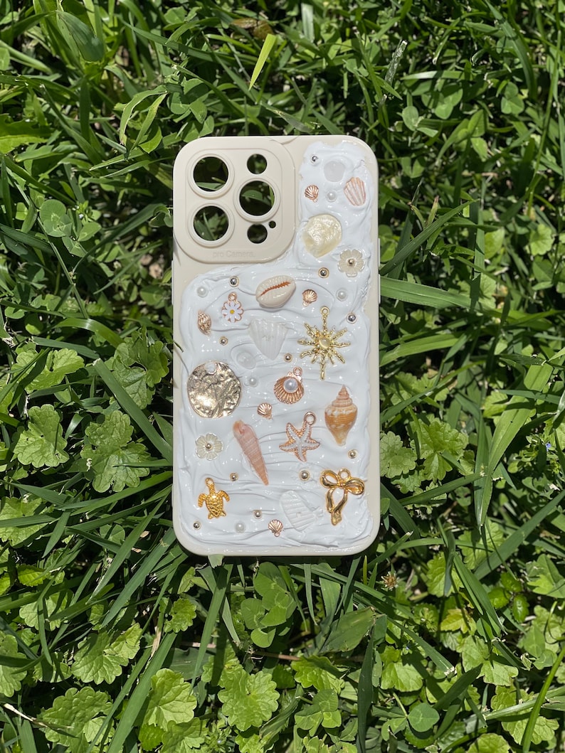 May include: A white iPhone case decorated with seashells, starfish, pearls, and gold accents. The case has a raised, textured design and a clear camera lens.