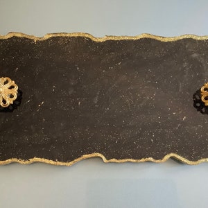 May include: Black serving tray with a gold glitter finish and a wavy edge. The tray has two gold metal decorative handles.