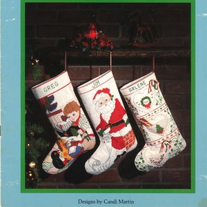 May include: A Christmas stocking collection with three stockings featuring cross stitch designs. The stockings are hung on a brick fireplace mantel. The stockings are decorated with a Santa Claus, a child, and a heart with a wreath. The stockings are labelled "Greg", "Jim", and "Arlene".