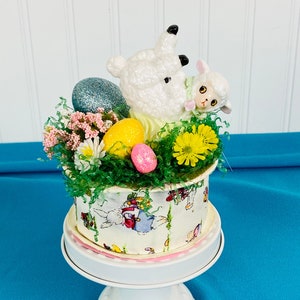 May include: A white ceramic Easter cake stand with a white and pink base. The stand is decorated with a white lamb figurine, a smaller lamb figurine, and pastel colored Easter eggs. The stand is surrounded by green grass and pink and yellow flowers.