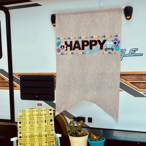 May include: A beige banner with the words "HAPPY Trails" and a peace sign, flowers, and a van graphic. A yellow and white lawn chair, potted plants, and a blue cooler sit on a blue patterned rug. The banner is hanging on the side of a camper.