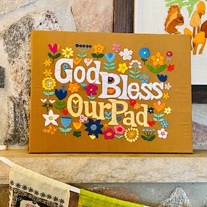 Könnte beinhalten: Eine braune Leinwand mit bunten gestickten Blumen und dem Text "God Bless Our Pad".