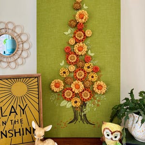 Puede incluir: Un tapiz de pared de tela verde con un diseño de árbol hecho de flores de tela naranja, amarilla y marrón. El tronco del árbol está bordado con hilo marrón. Una figura de ciervo, una figura de búho y un letrero enmarcado están en un estante debajo.