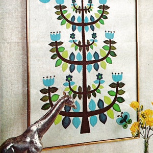 May include: A framed embroidery of a stylized tree with blue flowers and green leaves. The tree is brown and has a whimsical design. A silver giraffe figurine is in the foreground.