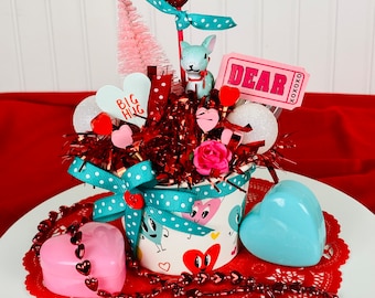 May include: A Valentine's Day themed centerpiece with a white ceramic cup with red hearts and a blue polka dot ribbon. The cup is filled with red and white tinsel, a pink and white miniature Christmas tree, a red glitter heart, a white bunny figurine, a pink ticket with the word "DEAR" on it, and a pink heart with the words "BIG HUG" on it. The centerpiece is on a white cake stand with a red lace doily and red heart beads.