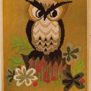May include: Framed needlepoint art featuring a detailed owl perched on a tree stump. The owl has large, expressive eyes and is surrounded by green leaves and red berries. The background is a textured gold color.
