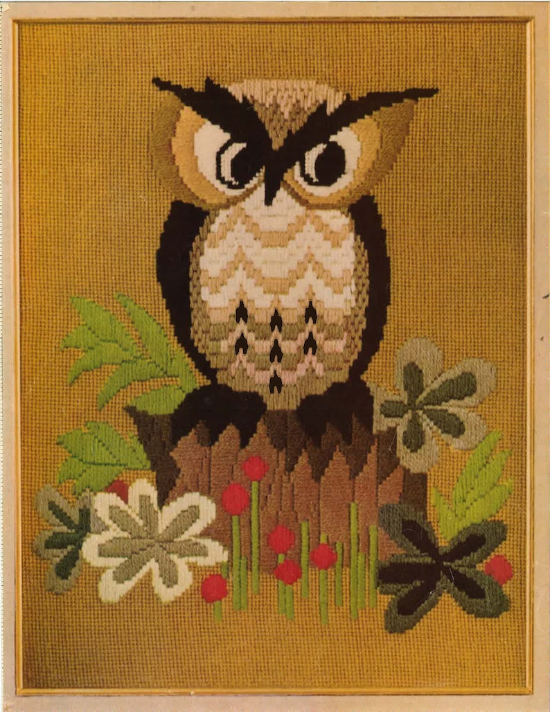 May include: Framed needlepoint art featuring a detailed owl perched on a tree stump. The owl has large, expressive eyes and is surrounded by green leaves and red berries. The background is a textured gold color.
