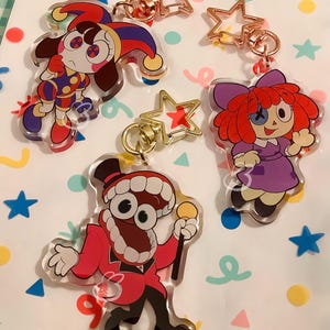 Puede incluir: Cuatro llaveros de personajes de dibujos animados. Un llavero presenta un buf&oacute;n en rojo, morado y amarillo. Otro muestra una mu&ntilde;eca con un vestido morado y pelo rojo. El tercer llavero es un personaje con un traje rojo. Cada llavero tiene un cierre en forma de estrella.
