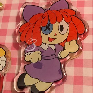 Puede incluir: Llavero acr&iacute;lico transparente de Raggedy Ann con el dise&ntilde;o cl&aacute;sico de la mu&ntilde;eca. La mu&ntilde;eca tiene pelo de hilo rojo, un lazo morado y un vestido morado. Un ojo es un bot&oacute;n con una X, el otro es un punto negro. El llavero tiene un cierre dorado.