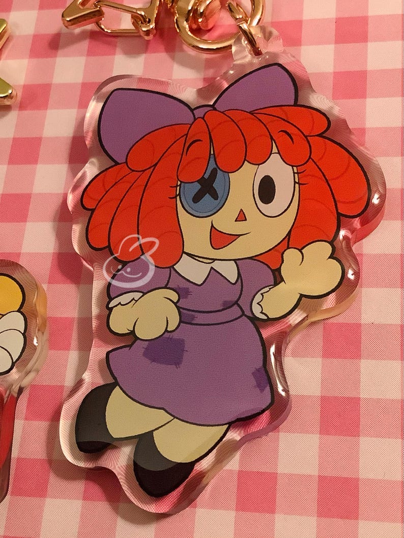 Puede incluir: Llavero acr&iacute;lico transparente de Raggedy Ann con el dise&ntilde;o cl&aacute;sico de la mu&ntilde;eca. La mu&ntilde;eca tiene pelo de hilo rojo, un lazo morado y un vestido morado. Un ojo es un bot&oacute;n con una X, el otro es un punto negro. El llavero tiene un cierre dorado.
