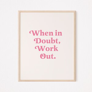 May include: A framed print with the text "When in Doubt, Work Out" in pink letters on a cream background. The frame is a light wood color, and the print is centered on a white wall.