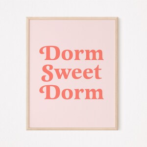 May include: A framed print with the words "Dorm Sweet Dorm" in coral-colored, bold, retro-style font. The text is centered on a light pink background, and the frame is a light wood color.