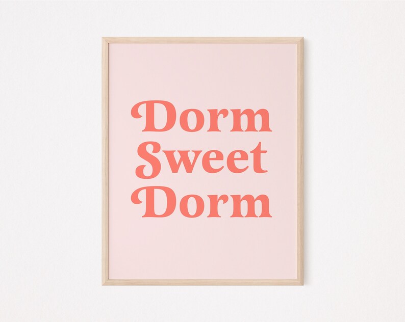 May include: A framed print with the words "Dorm Sweet Dorm" in coral-colored, bold, retro-style font. The text is centered on a light pink background, and the frame is a light wood color.