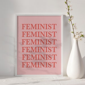 Puede incluir: Una impresi&oacute;n art&iacute;stica enmarcada con la palabra "FEMINIST" repetida en color coral sobre un fondo rosa claro. Un jarr&oacute;n blanco con flores blancas se encuentra a la derecha de la impresi&oacute;n. La impresi&oacute;n se muestra sobre una superficie blanca.