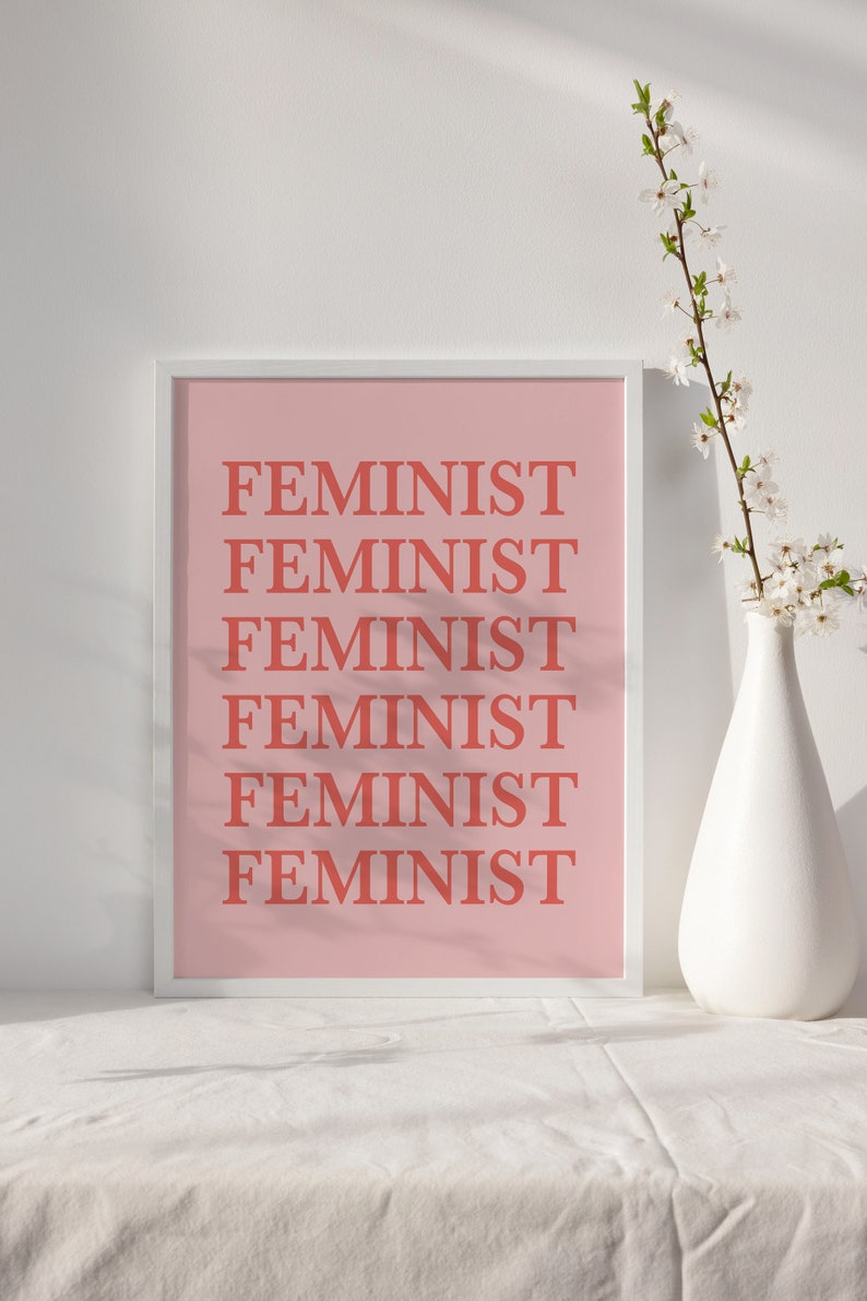 Puede incluir: Una impresi&oacute;n art&iacute;stica enmarcada con la palabra "FEMINIST" repetida en color coral sobre un fondo rosa claro. Un jarr&oacute;n blanco con flores blancas se encuentra a la derecha de la impresi&oacute;n. La impresi&oacute;n se muestra sobre una superficie blanca.