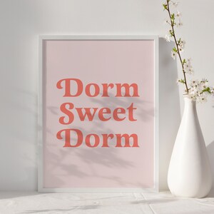 May include: A framed print with the words "Dorm Sweet Dorm" in coral-colored, retro-style lettering on a light pink background. A white vase with white flowers sits to the right of the framed print. The print is on a white surface.